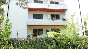 A multi-story building with a white facade, balconies with plants, and a red roof.