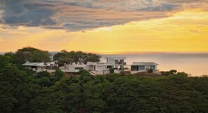 A modern house with multiple levels, surrounded by dense greenery and overlooking a body of water during sunset.