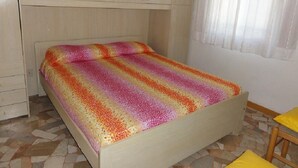A small, single-bed room with a plain wooden headboard, a patterned bedspread, and a tiled floor.