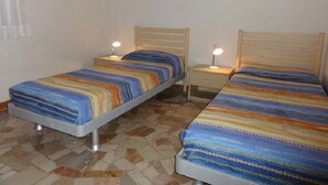 A room with two single beds, each with a wooden headboard and a bedside table with a lamp. The floor is tiled with a mosaic pattern.