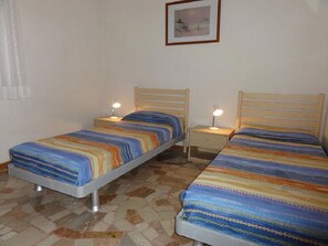 Quarto com duas camas individuais, cada uma com cabeceira de madeira e mesa de cabeceira com abajur. O chão é revestido com um padrão de mosaico.