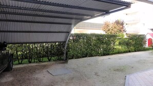 A covered outdoor area with a gravel ground, a metal roof, and a hedge in the background.