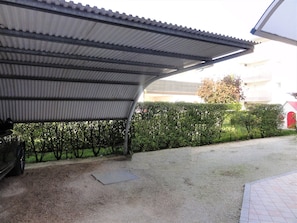 A covered outdoor area with a gravel ground, a metal roof, and a hedge in the background.