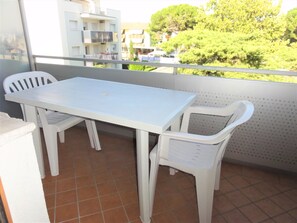 Uma varanda com mesa e cadeiras brancas, piso de azulejos e vista para edifícios e árvores.