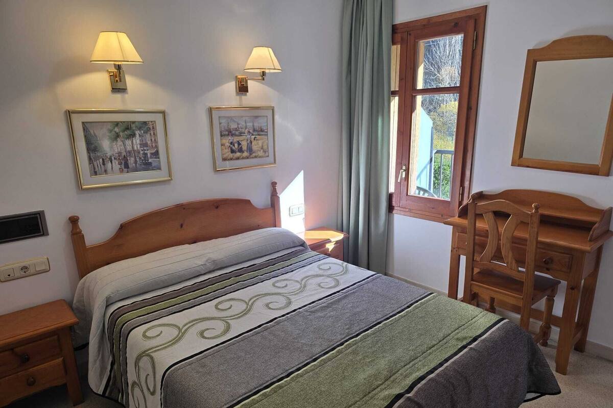 A bedroom with a bed, two wall-mounted lamps, a dresser, a window with curtains, and two framed pictures on the wall.