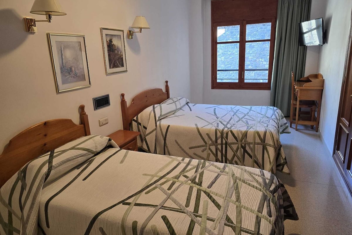 A hotel room with two beds, a window with curtains, a TV, and wall-mounted lamps.