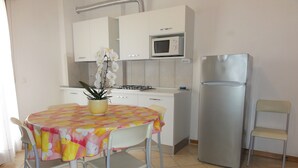 Une petite cuisine avec des meubles blancs, un four à micro-ondes, un réfrigérateur, une table à manger recouverte d’une nappe colorée et un pot d’orchidée.