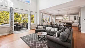 A spacious living room with a gray sectional sofa, a round coffee table, and a patterned rug. There are large windows with curtains, a dining area, and a kitchen in the background. - Modern Vacation Home w/ Private Pool (Hilton Head Island)