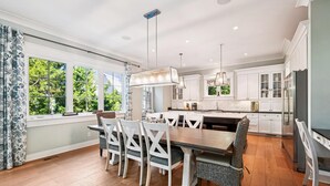 A modern kitchen with white cabinetry, a central island, and a dining area with white chairs. - Modern Vacation Home w/ Private Pool (Hilton Head Island)