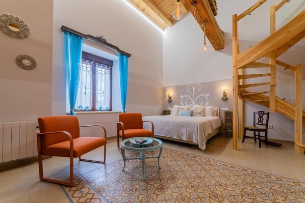 Un dormitorio con cama, dos sillas naranjas, una mesita, una ventana con cortinas azules, una escalera de madera y apliques decorativos.