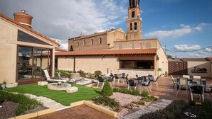 Un patio con mobiliario exterior, una torre de campanas y un edificio con grandes ventanales.