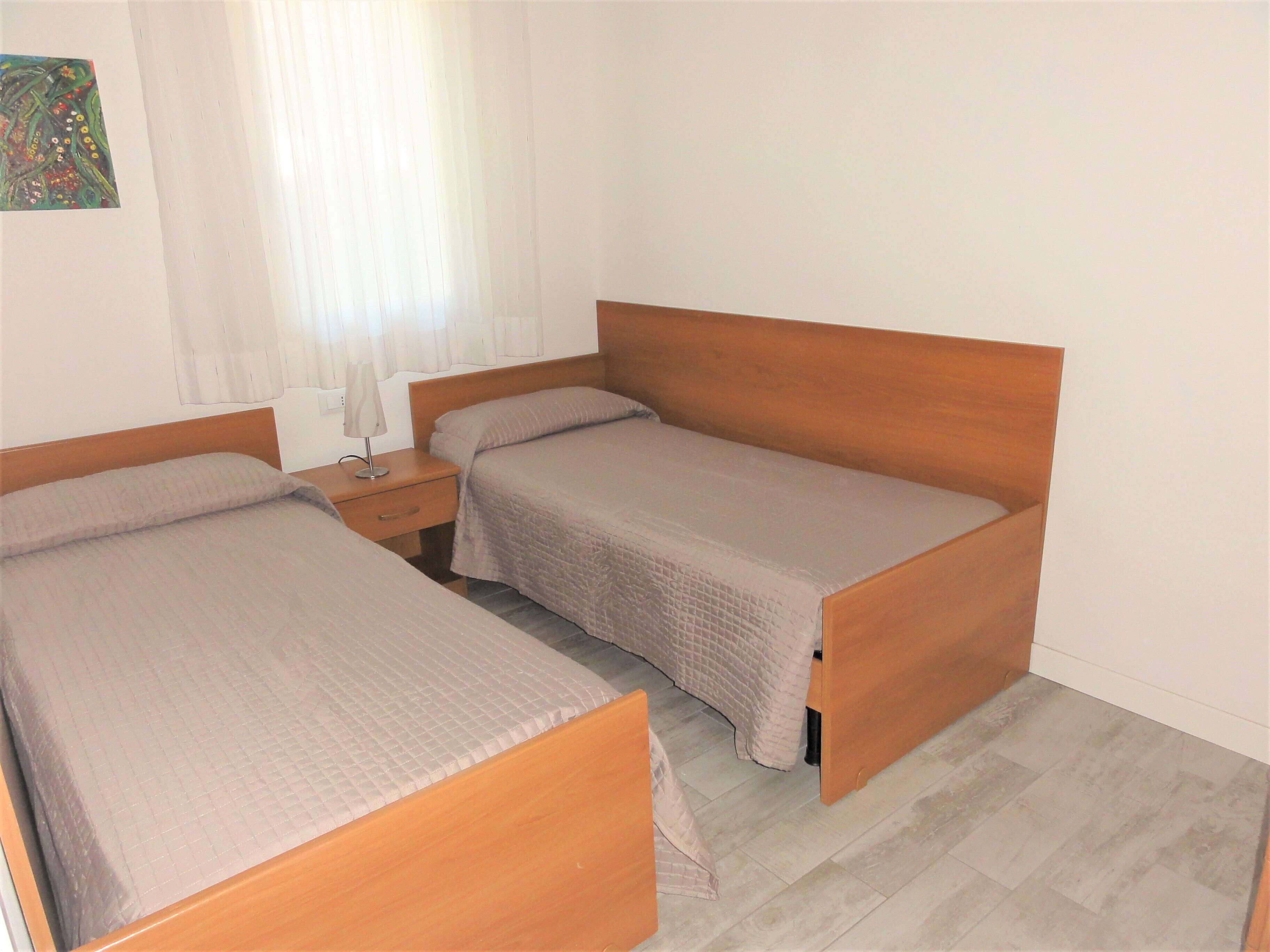A room with two single beds, a nightstand with a lamp, and a window with curtains.