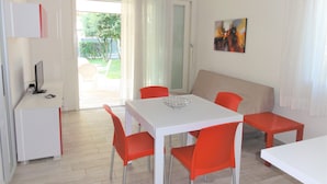 A modern living room with a white sofa, a dining table with four chairs, and a TV on a stand. - Villa Only 100mt From the Beach (San Michele al Tagliamento)
