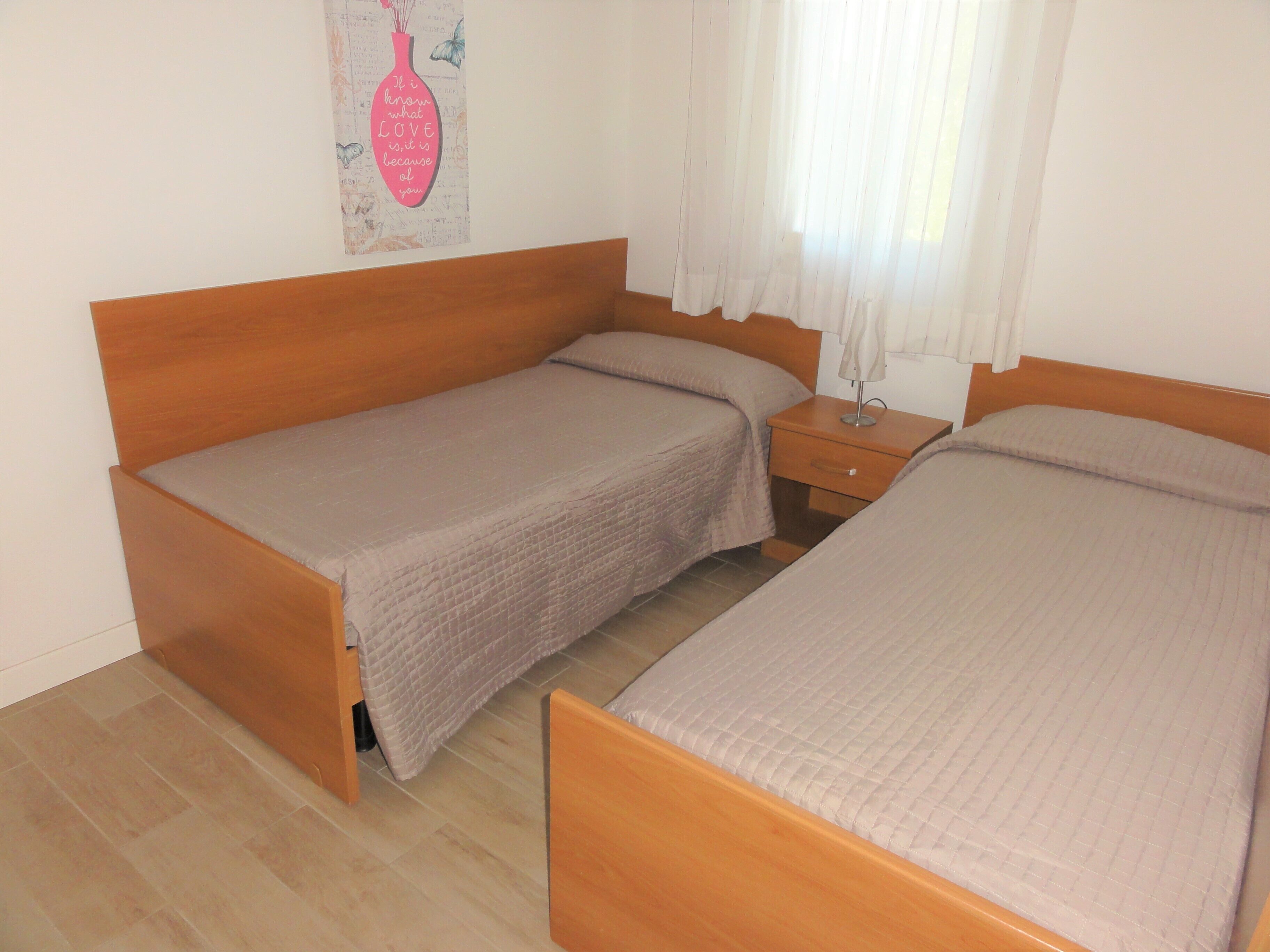 Ein Schlafzimmer mit zwei Holzbett, einem Nachttisch, einer Wanddekoration und einem Fenster mit leichtem Vorhang.