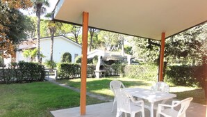 Une terrasse extérieure avec une table et des chaises, entourée d’un jardin et avec une maison en arrière-plan.