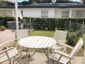 Un patio con una mesa redonda y cuatro sillas, un edificio blanco con techo de tejas, y un seto al fondo.