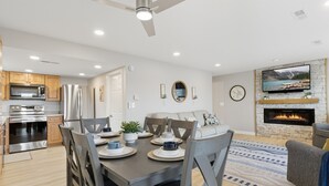 Eine moderne Küche mit Edelstahlgeräten, ein Essbereich mit Tisch für vier Personen und ein Wohnzimmer mit Kamin und Wandfernseher.