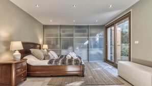 A modern bedroom with a large bed, a nightstand, a bench, and a sliding glass door leading to a balcony. - Charming Chalet | 168m2 | 6 pax | Familial | View (Les Houches)