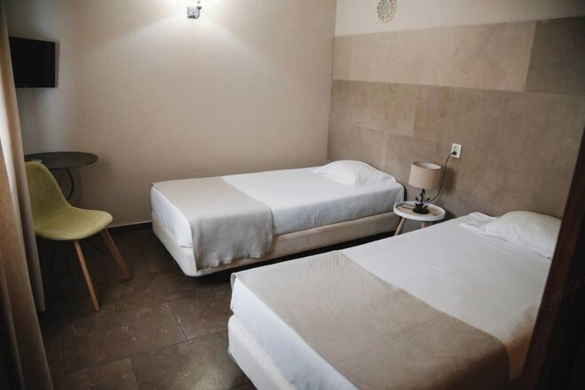 A hotel room with two single beds, a small table with a lamp, a chair, and a television mounted on the wall.
