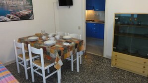 A dining area with a table set for four, white chairs, a television mounted on the wall, and a kitchen visible through an open doorway.