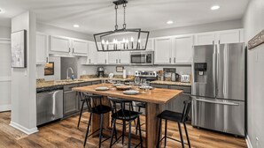 Une cuisine moderne avec des meubles blancs, des appareils en acier inoxydable, un îlot central en bois doté de tabourets, et un lustre.
