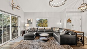 Un salon moderne avec un canapé d’angle, une table basse ronde et un grand tapis. Il y a deux fenêtres donnant sur des arbres et une porte ornée d’un élément décoratif mural.