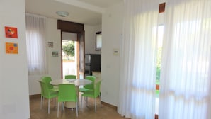 A dining area with a round table and green chairs, a kitchen area with a refrigerator, and a view of greenery outside. - Comfy Villa With Fenced Garden Near the Beach (San Michele al Tagliamento)