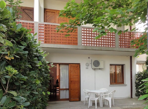 Un bâtiment à deux étages doté d’un balcon en bois, d’une porte blanche et d’un petit espace extérieur aménagé avec des chaises en plastique.