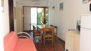 A compact room with a sofa, a dining table with chairs, a small kitchen area with a refrigerator, and a window with a view of greenery. - Flat in a Villa With Private Garden (San Michele al Tagliamento)