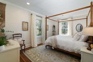 A four-poster bed with a wooden frame, a white bedspread, and matching pillows. There is a wooden chair with a cushion, a small table with a lamp, and a decorative mirror on the wall. - The Potters House: Luxury Apartment/Walk Downtown (Staunton)