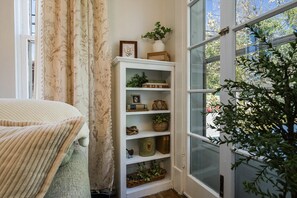 Una esquina de una habitación con un estante blanco que contiene libros, objetos decorativos y una planta. Se ve una ventana con vistas a árboles y un coche estacionado.
