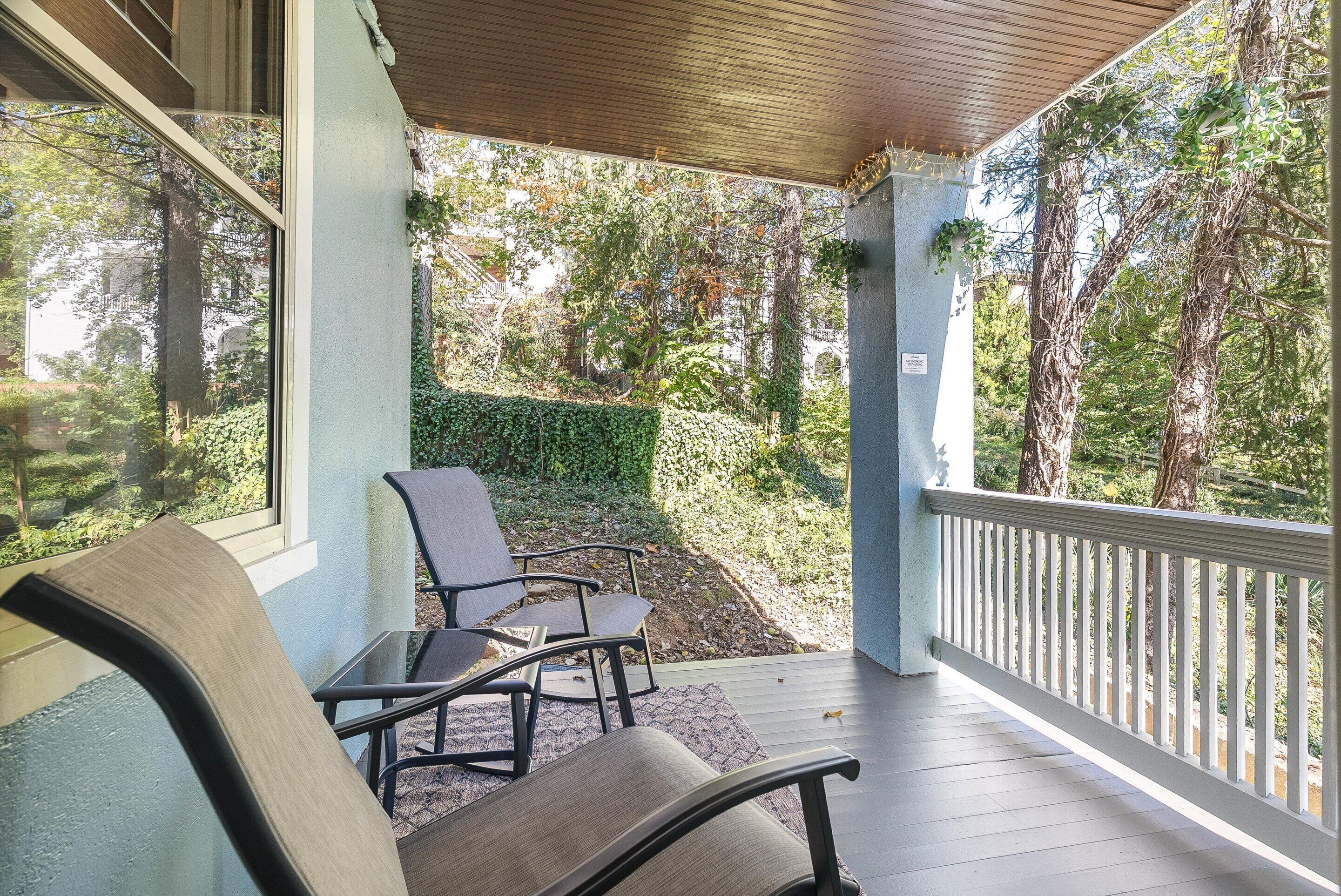 A covered outdoor seating area with two chairs and a table, overlooking a garden with trees and a hedge.