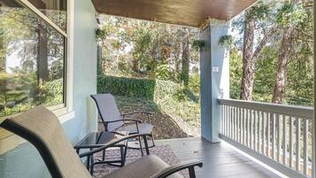 A covered outdoor seating area with two chairs and a table, overlooking a garden with trees and a hedge.