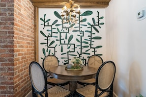 A round dining table with four wicker chairs, a chandelier, and a decorative wall with leaf patterns. - Jungalu: Lush Jungle Retreat Downtown (Staunton)