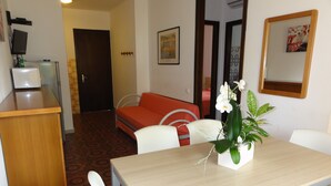 Quarto de hotel com área de refeições, sofá, mesa pequena com planta e televisão em um suporte.