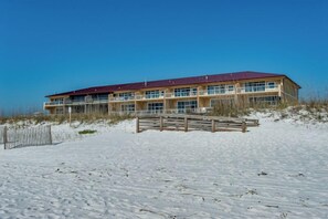 Un edificio grande de varios pisos con techo rojo, ubicado en una playa de arena con una valla de madera al frente.