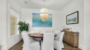 Une salle à manger avec une table ronde en bois, des chaises blanches, un lustre, un tableau représentant une vague et un meuble contenant des objets décoratifs.
