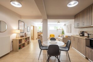 A modern kitchen with wooden cabinets, a dining area with a round table and chairs, and a bright, well-lit living space with a sofa and a yellow chair.