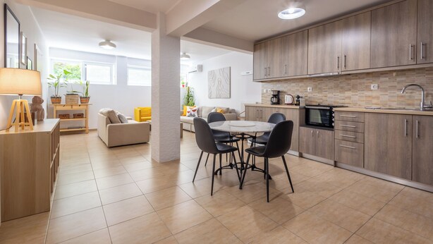 Een moderne keuken met houten kastjes, een eetgedeelte met een ronde tafel en stoelen, en een zithoek met een bank en sierplanten.