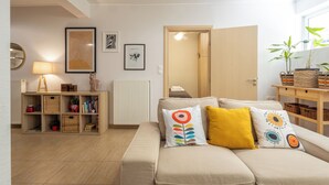 Een gezellige woonkamer met een bank, kussens, een boekenkast, een lamp en een deur naar een andere kamer.