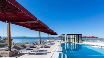 Un espace piscine aménagé sur le toit, avec des chaises longues et des parasols rouges, offrant une vue sur un ciel bleu dégagé et un paysage lointain.