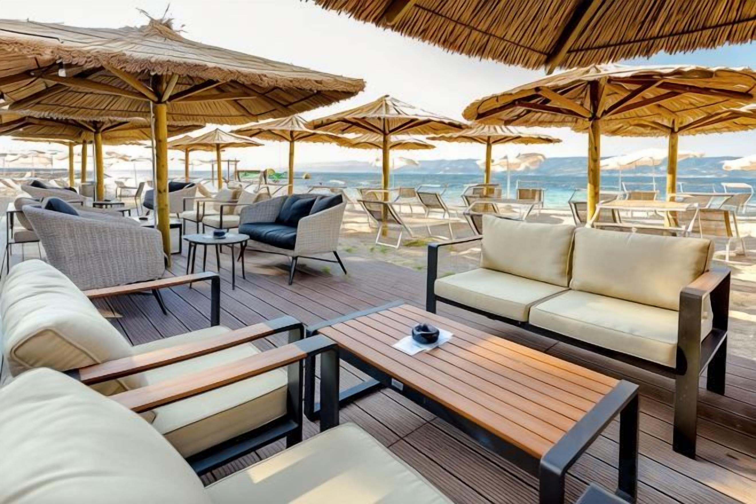 An outdoor seating area with wooden tables and cushioned chairs, surrounded by thatched umbrellas, overlooking a beach.