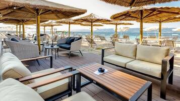 An outdoor seating area with wooden tables and cushioned chairs, surrounded by thatched umbrellas, overlooking a beach.
