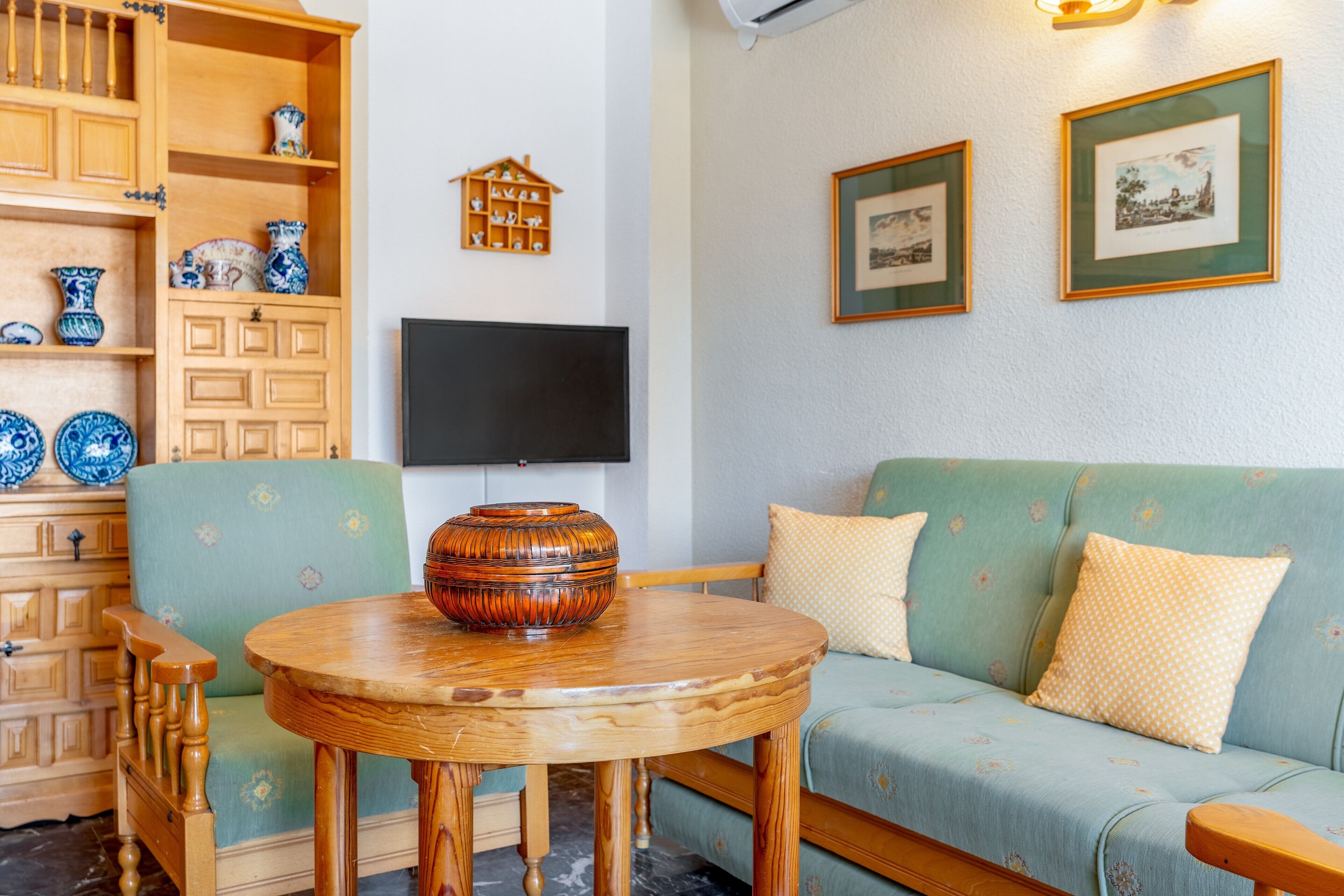 Una sala de estar con un sofá verde, una mesa de madera con un florero, un televisor y un mueble de madera con objetos azules y blancos.