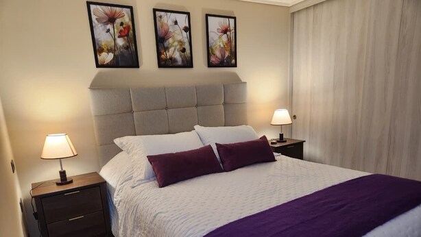 A neatly made bed with a white and purple bedspread, two bedside lamps, and three floral wall art pieces above the bed. - Fully equipped apartment, bedroom en suite in Valdivia, direct access to the city entrance. (Valdivia)