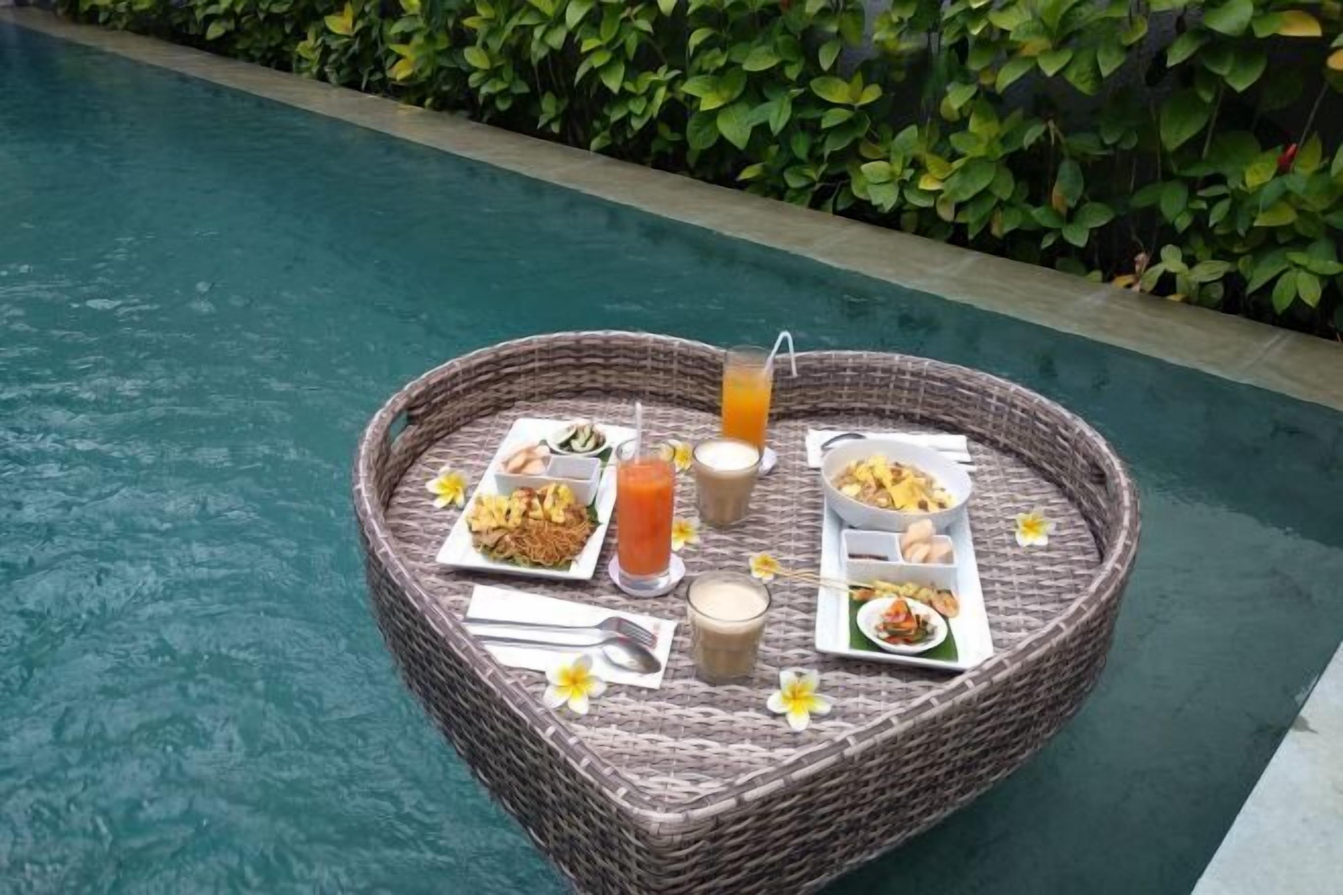 Een rieten tafel met een ontbijtbuffet bij een zwembad, inclusief borden met eten, drankjes en een glas sinaasappelsap.