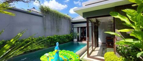 A swimming pool with a water feature and a floating platform, surrounded by greenery and a modern house.
