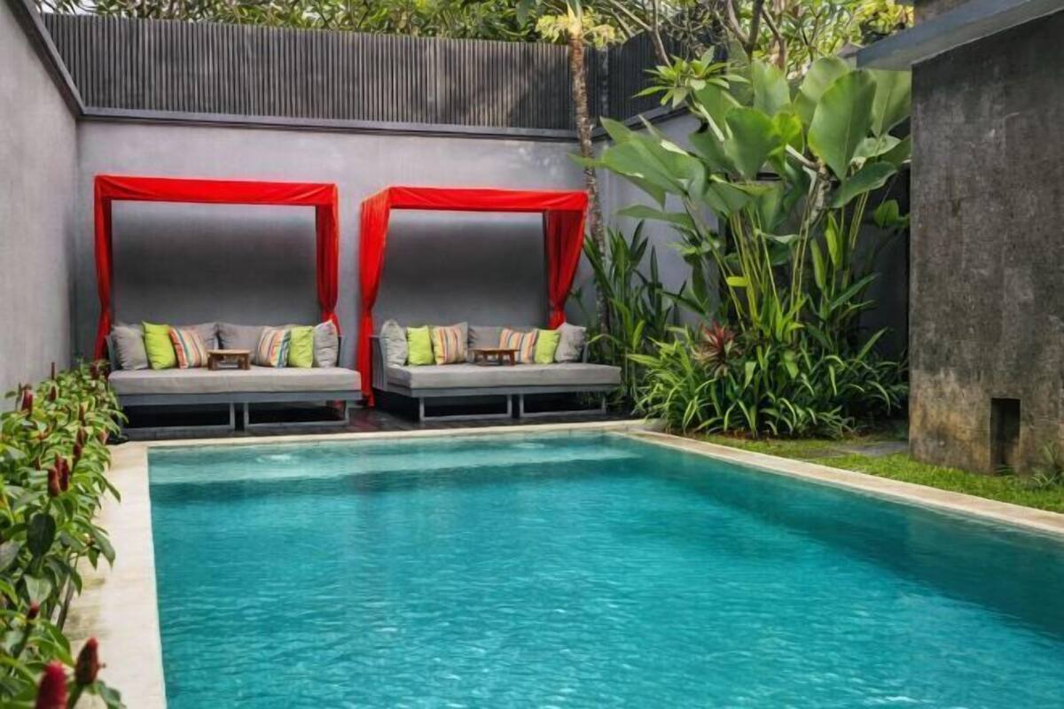 A pool area with two red canopies, grey lounge chairs with cushions, and surrounding greenery.