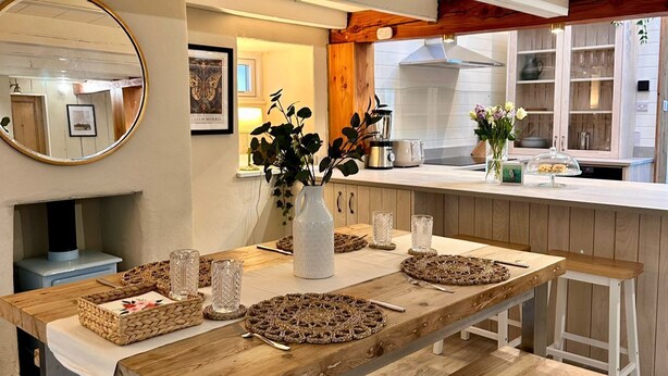 A modern kitchen with a wooden dining table, benches, and a central island. - Pilgrim Cottage - sleeps 4 guests  in 2 bedrooms (Newquay)