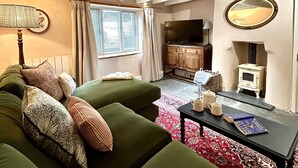 A cozy living room with a green sofa, patterned cushions, a small table with books and mugs, a fireplace, and a television. - Pilgrim Cottage - sleeps 4 guests  in 2 bedrooms (Newquay)